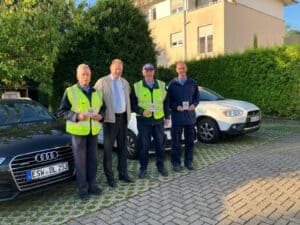 Vier Männer vor Autos, zwei tragen Westen.